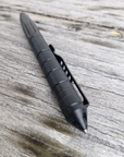 The Op Gear Tactical Pen - Recon, featuring a black metal body, textured grip, and clip, lies diagonally on a weathered wooden surface—combining sleek design with self-defence functionality.