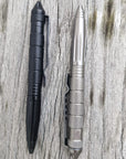 Two Op Gear Tactical Pen - Recon pens, one black and one silver, rest side by side on weathered wood. Each pen features a textured grip and clip, serving as both a practical self-defense tool and a reliable writing instrument.