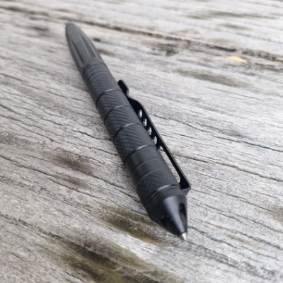 The Op Gear Tactical Pen - Recon, featuring a black metal body, textured grip, and clip, lies diagonally on a weathered wooden surface—combining sleek design with self-defence functionality.