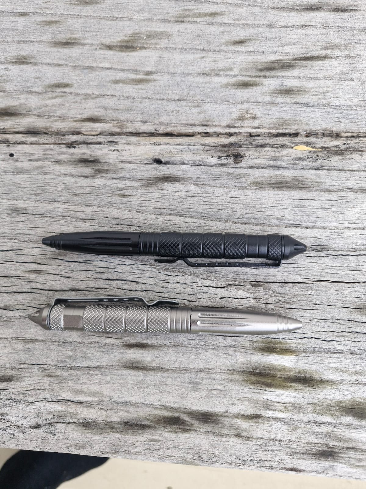Two Op Gear Tactical Pen - Recon models, one black and one silver, lie parallel on a weathered wooden surface. Both feature textured grips and pointed ends for dependable self-defense in daily use.